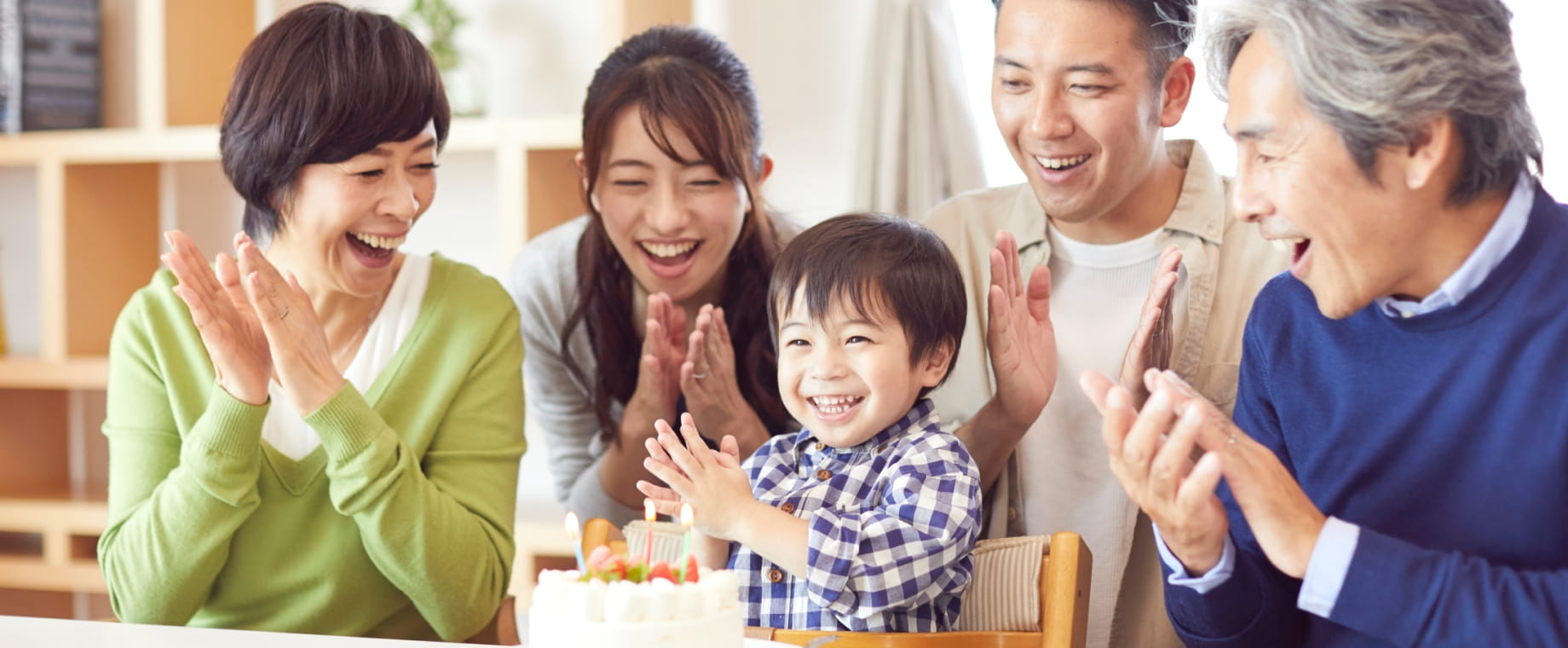 三世代家族（祖父母、若い両親、幼稚園くらいの男の子）が笑顔で誕生日ケーキを囲んで拍手している写真。