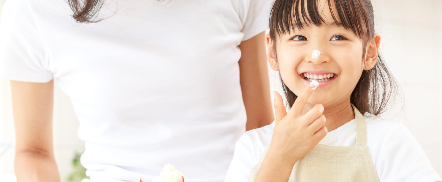 若い女性と小学生くらいの女の子がケーキを作っているシーン。女の子が笑顔でクリームを指につけて食べようとしている。